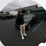 Person in dark clothing standing in a parking lot with parked vehicles in the background